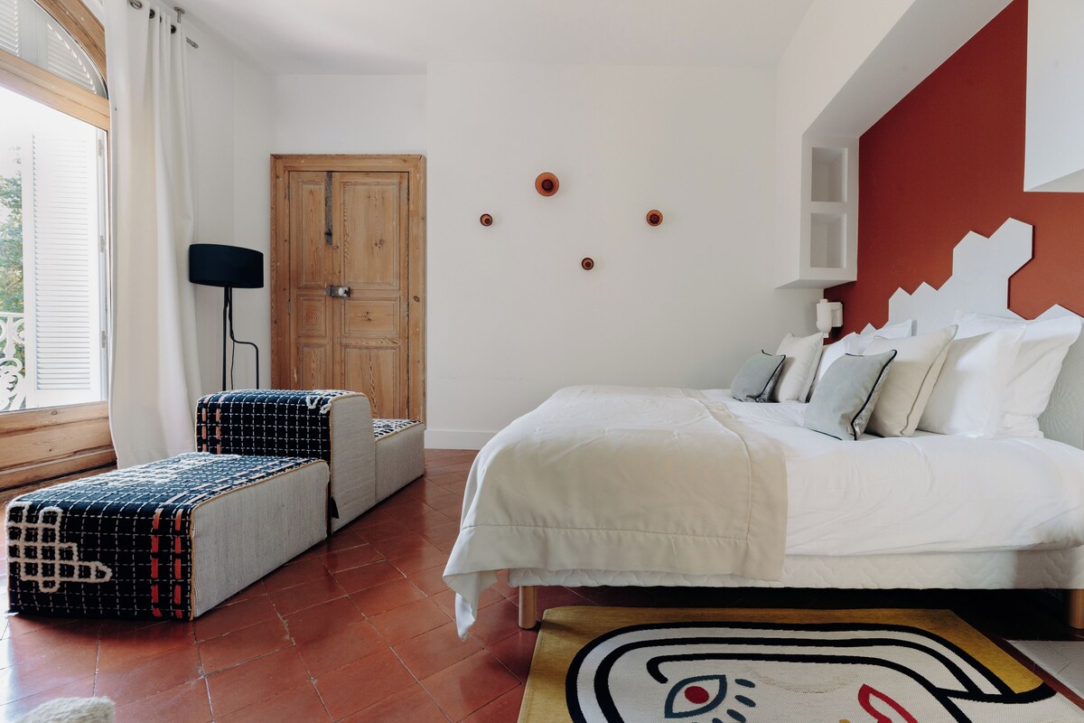 Un dormitorio con cama, una puerta de madera, una ventana con cortinas y una lámpara fijada a la pared.