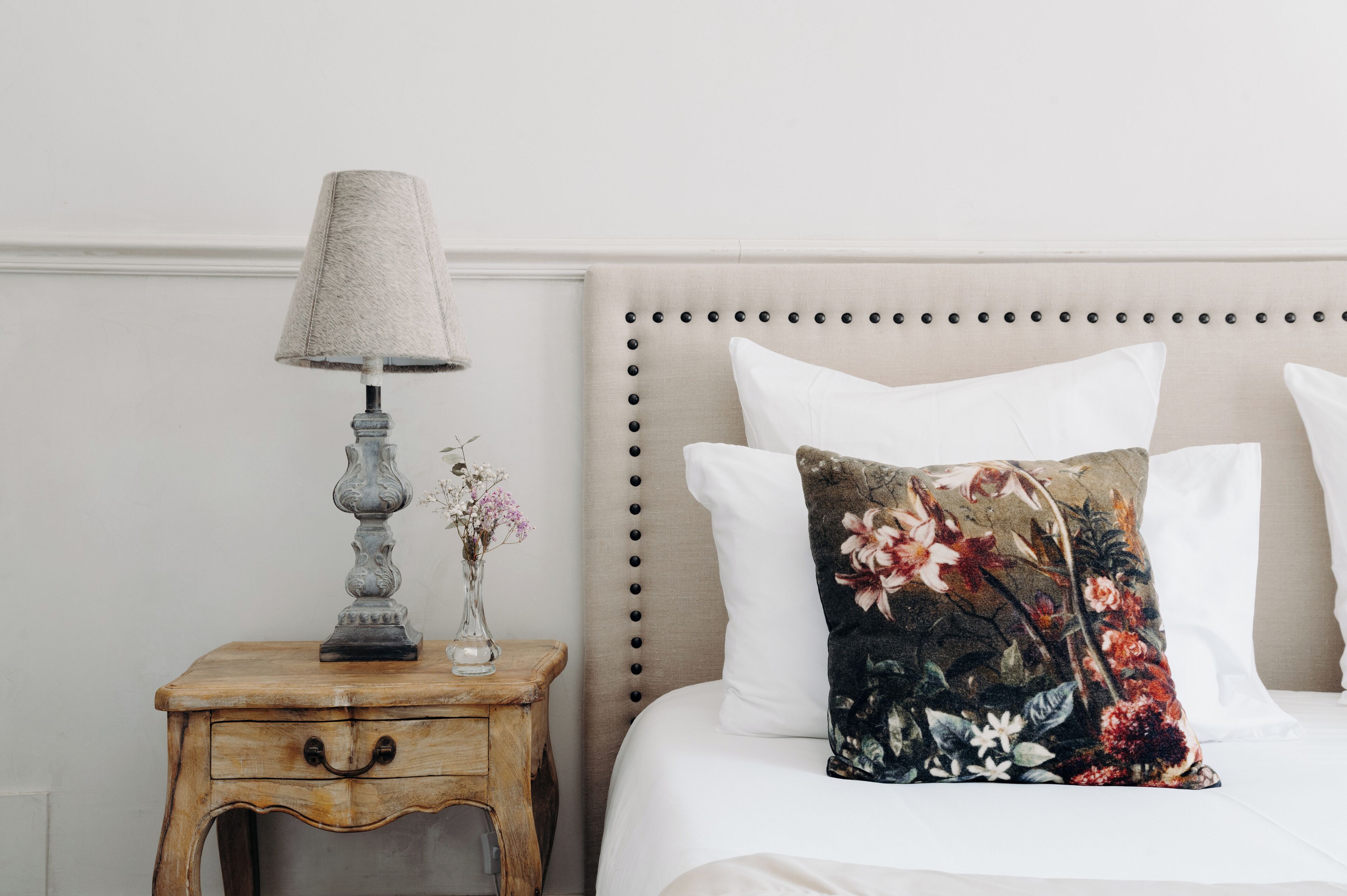 A bed with a floral pillow, a bedside lamp, and a small vase with flowers on a wooden nightstand.
