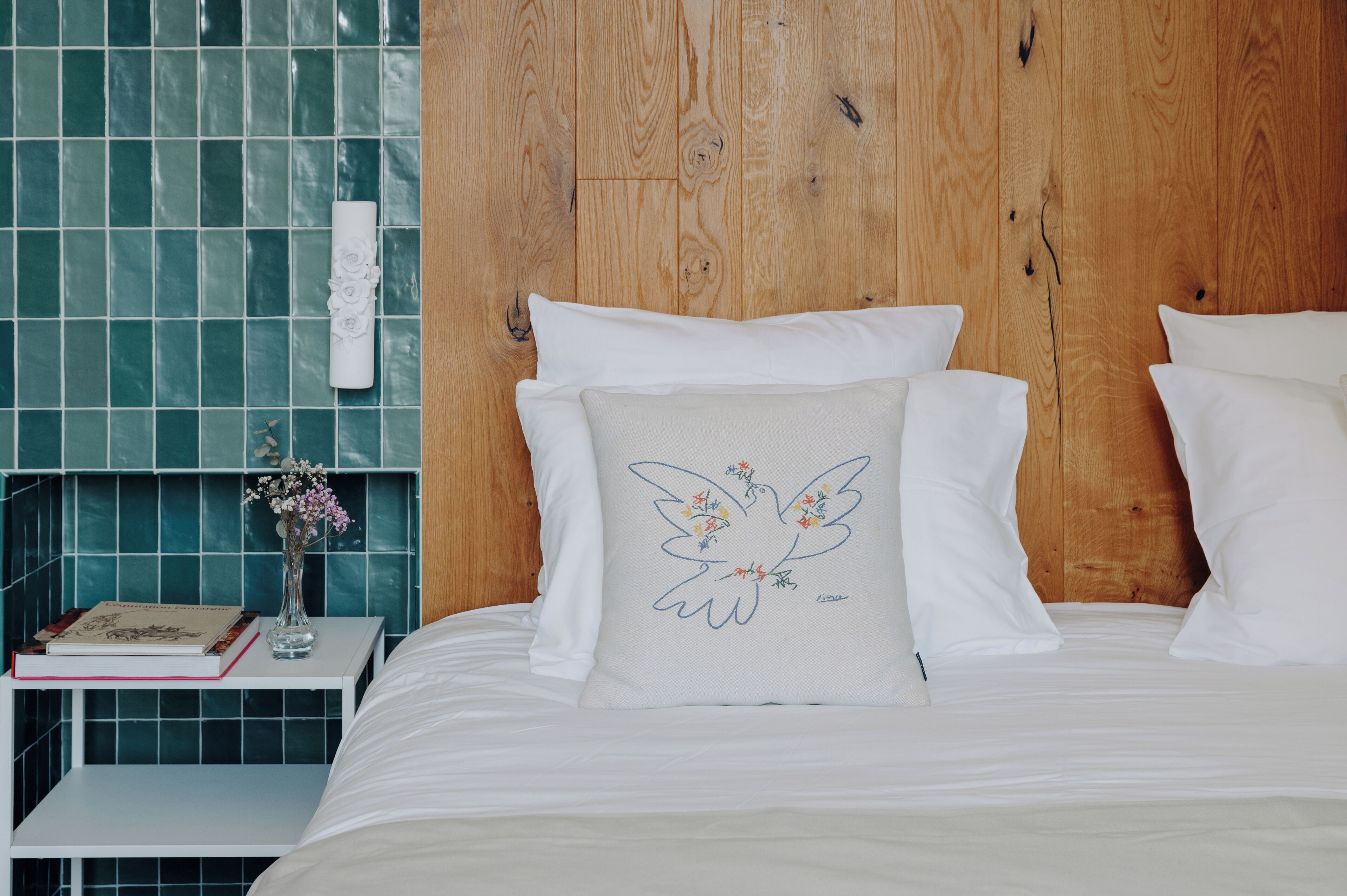 Una cama bien hecha con ropa de cama blanca y un cojín con un diseño de pájaro. Una mesita de noche sostiene un libro y un jarrón con flores. La habitación tiene un cabecero de madera y paredes con azulejos color aguamarina.