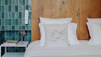 Una cama bien hecha con ropa de cama blanca y un cojín con un diseño de pájaro. Una mesita de noche sostiene un libro y un jarrón con flores. La habitación tiene un cabecero de madera y paredes con azulejos color aguamarina.