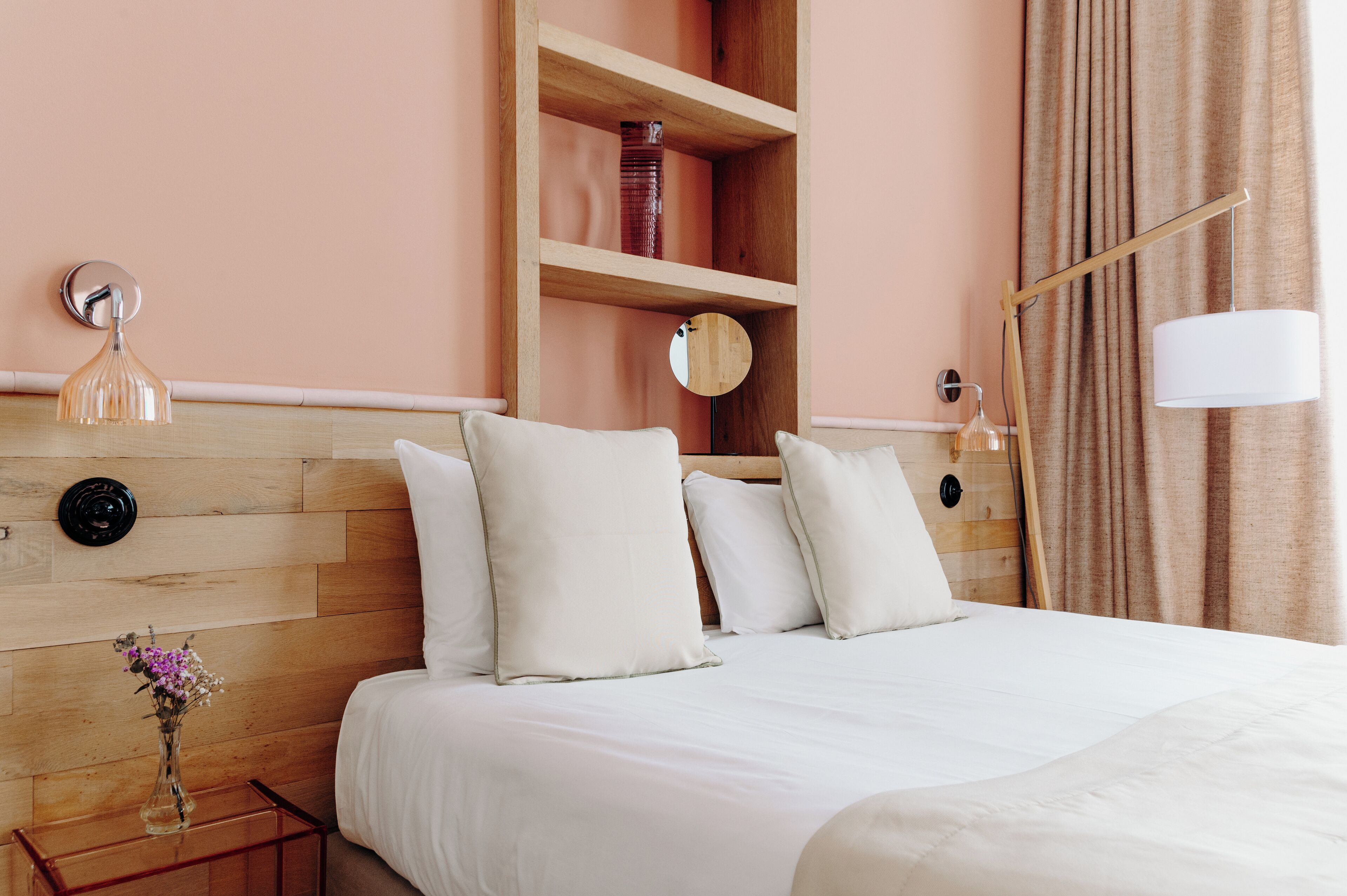 A neatly made bed with white linens and pillows, a wooden headboard, a bedside table with a vase of flowers, and a wall-mounted mirror.