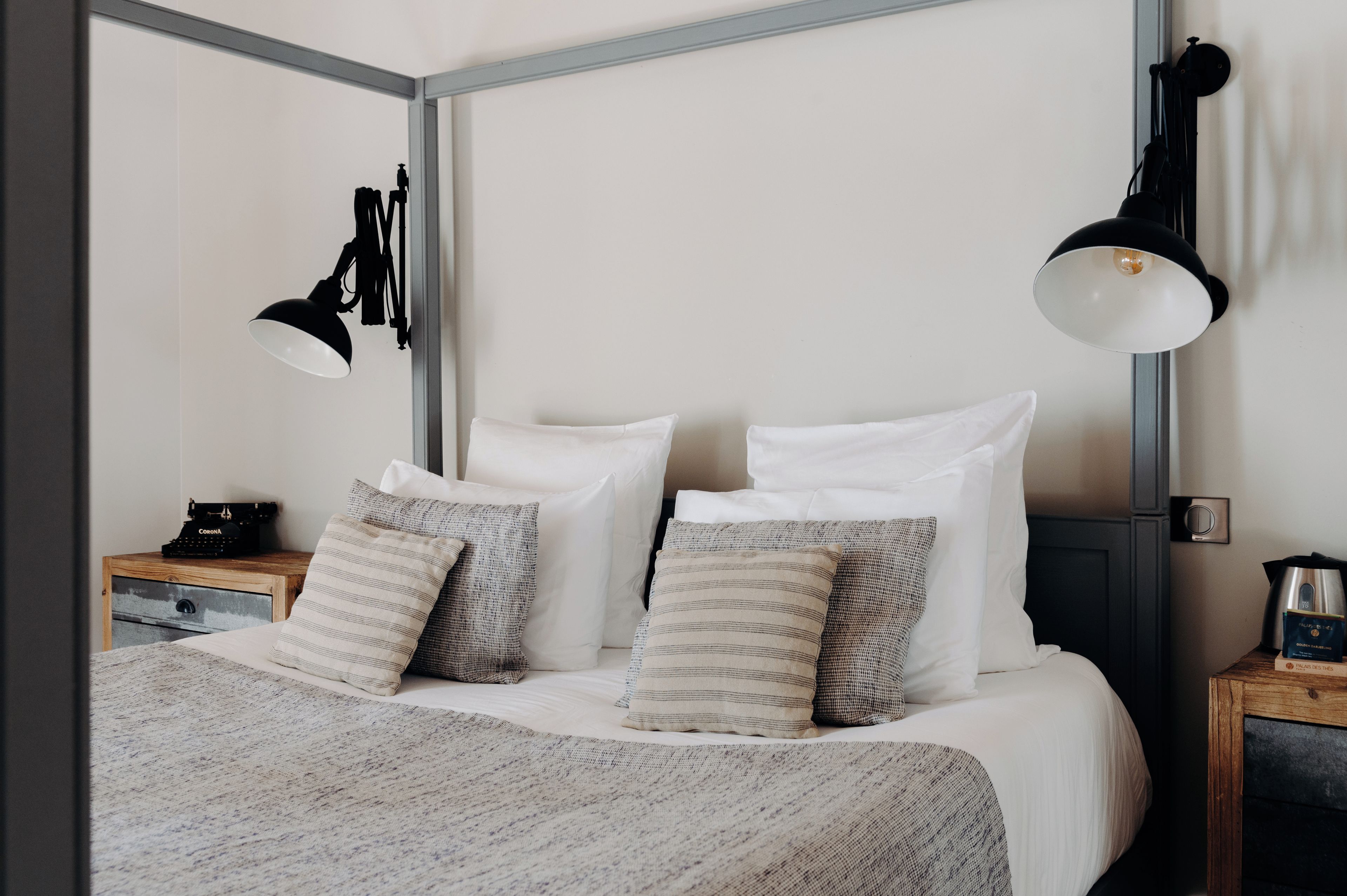 A neatly made bed with white and striped pillows, a wooden nightstand with a vintage typewriter, and a wall-mounted lamp.