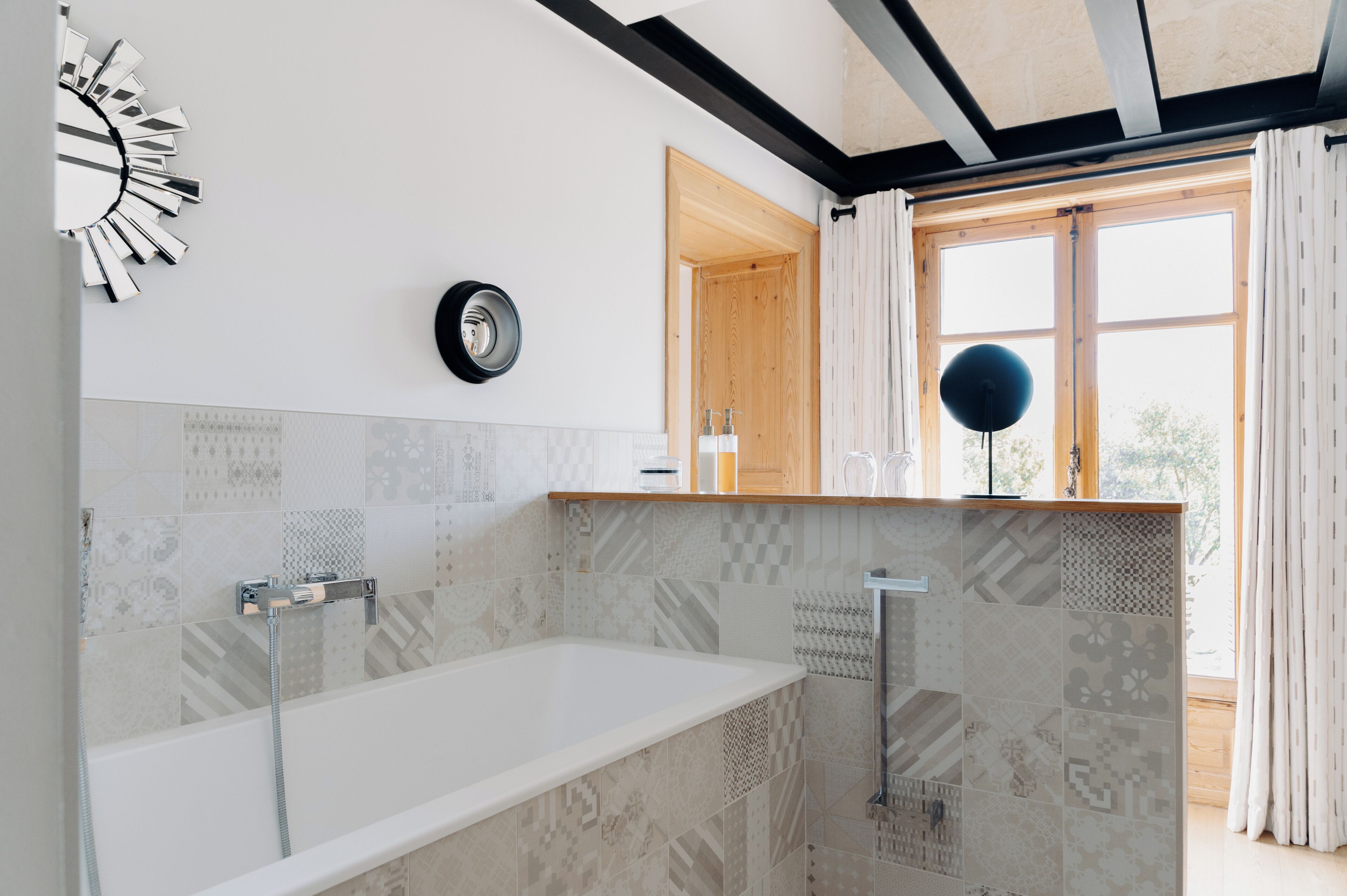 Un baño moderno con una bañera independiente, un espejo redondo y un ventanal con cortinas.