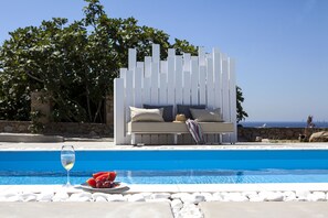 A poolside area with a white outdoor seating area, a glass of water, and a plate of watermelon and grapes.