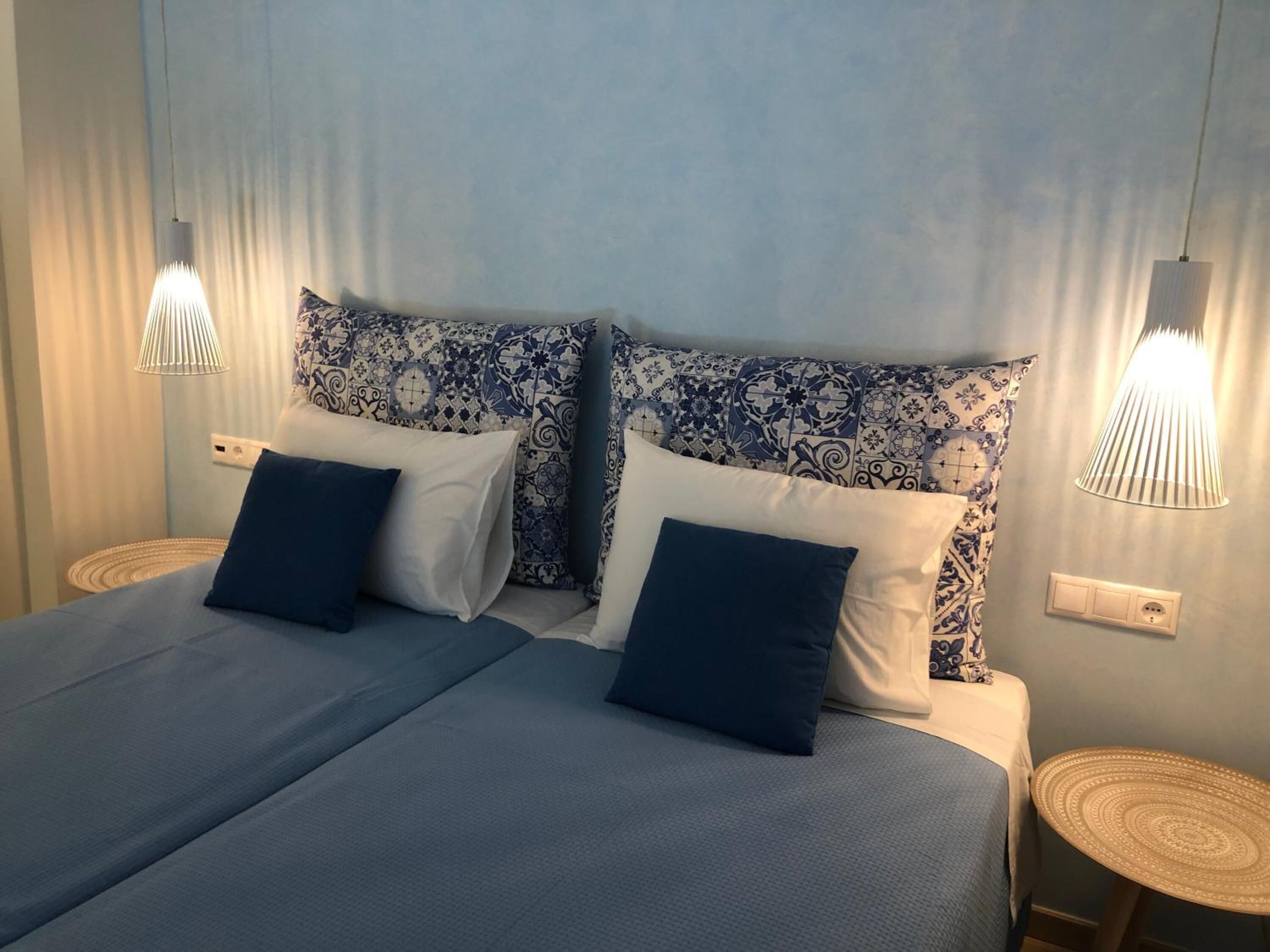 A double bed with blue bedding and patterned pillows, two bedside tables with lamps, and a light blue wall with a curtain.