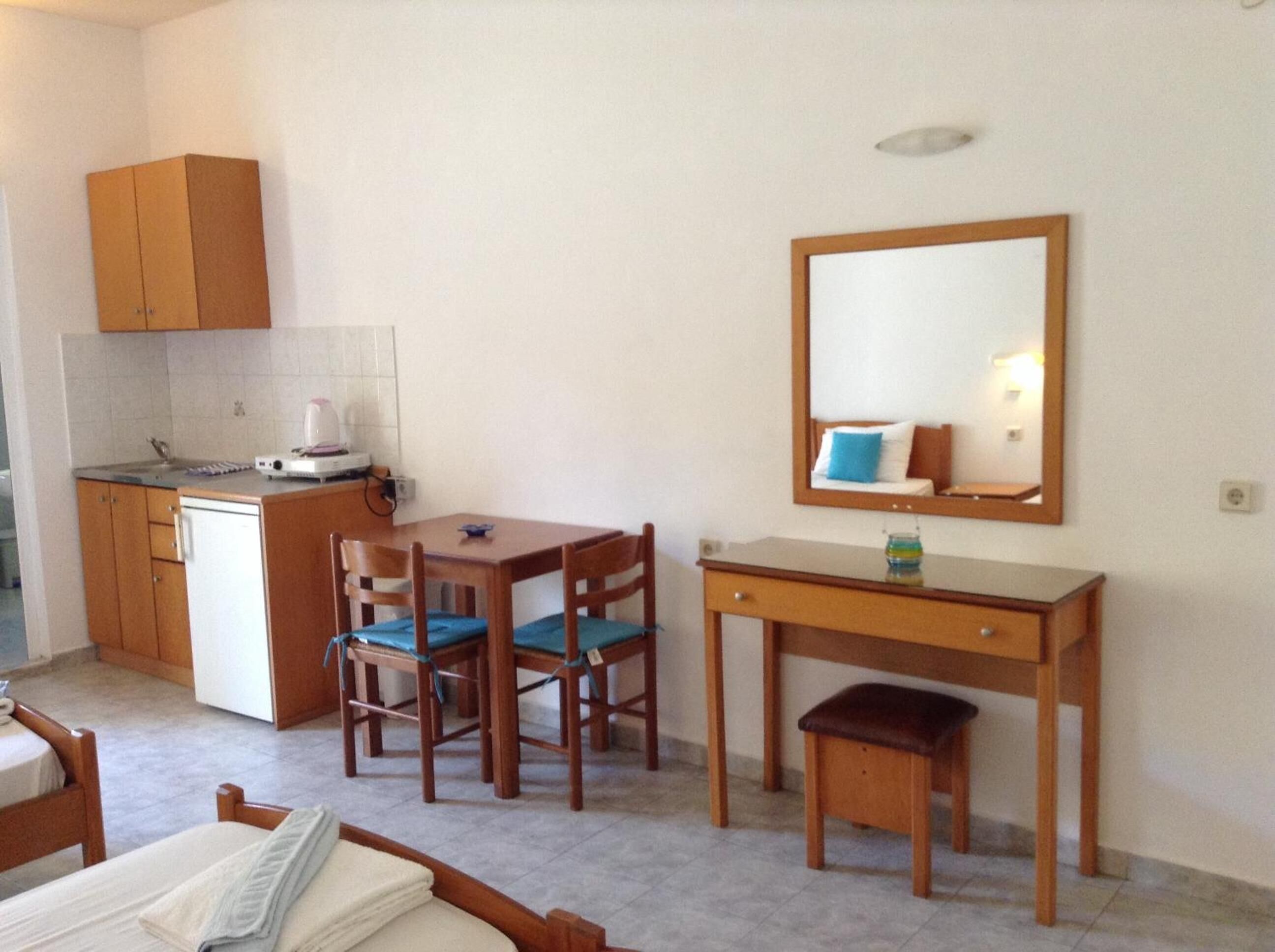 A compact room with a kitchenette, a dining area with chairs, a small desk, and a bed.