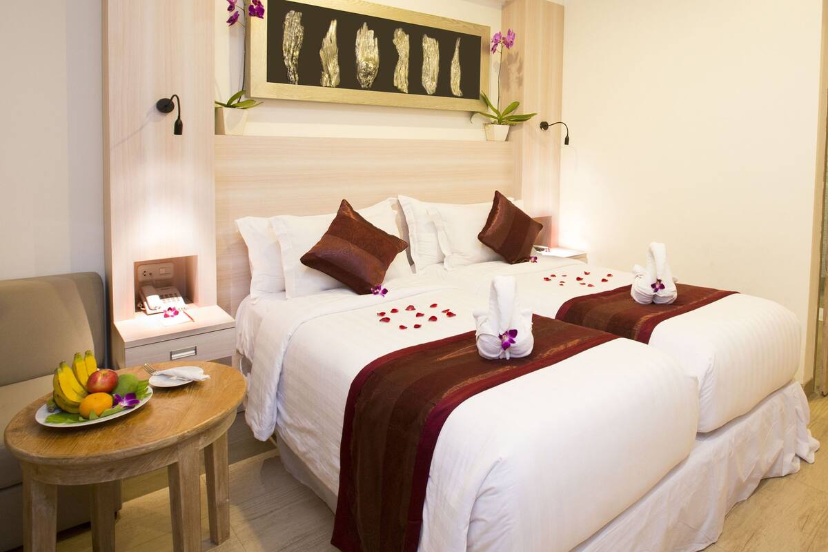 Habitación de hotel con dos camas, una mesita de noche con fruta, una pequeña mesa con un plato de fruta y un cuadro en la pared.