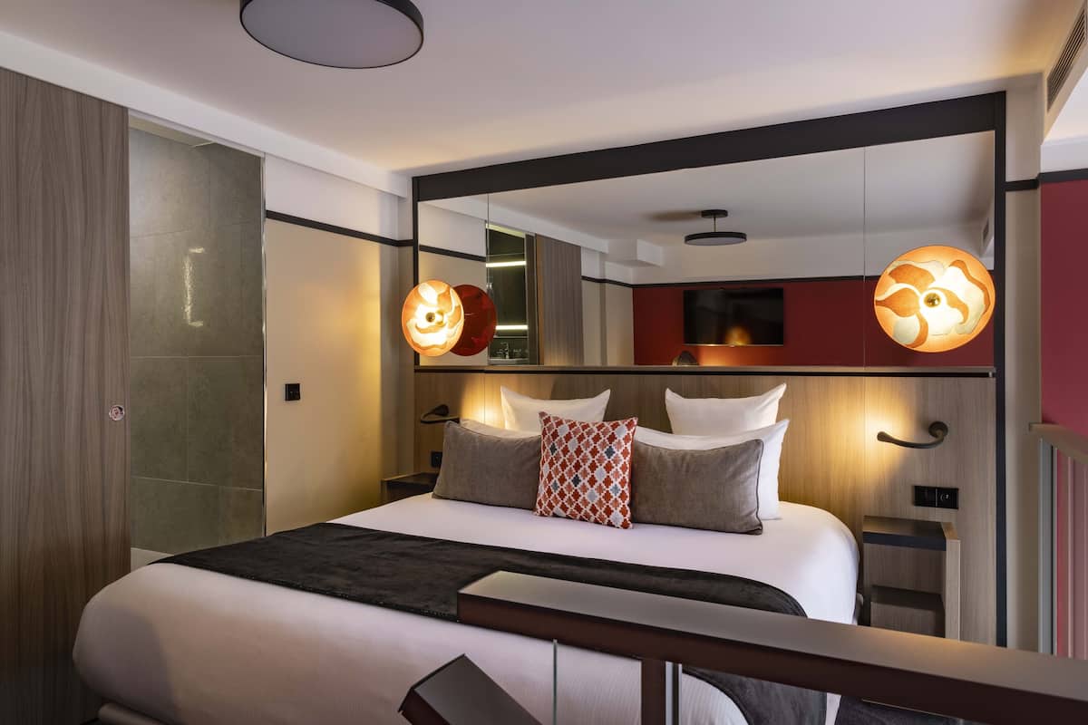 Una habitación de hotel moderna con una cama grande, un espejo sobre la cama y una mesita de noche con una lámpara.