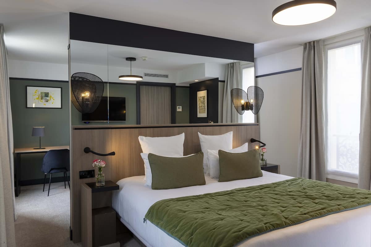 Una habitación de hotel moderna con una cama grande, un escritorio y una silla. Hay un espejo sobre la cama y una ventana con cortinas.