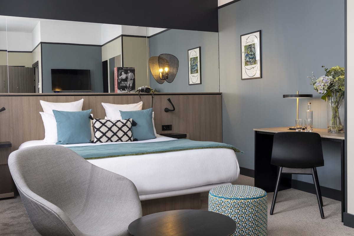 Una habitación de hotel moderna con una cama grande, un escritorio y una silla. Hay un espejo en la pared, un televisor de pantalla plana y un florero con flores.