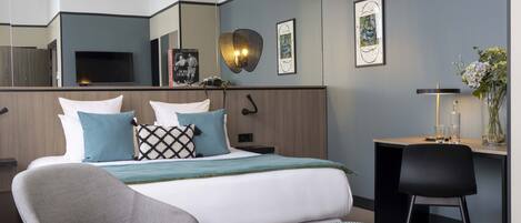 A modern hotel room with a large bed, a desk, and a chair. There is a mirror on the wall, a flat-screen TV, and a vase with flowers.