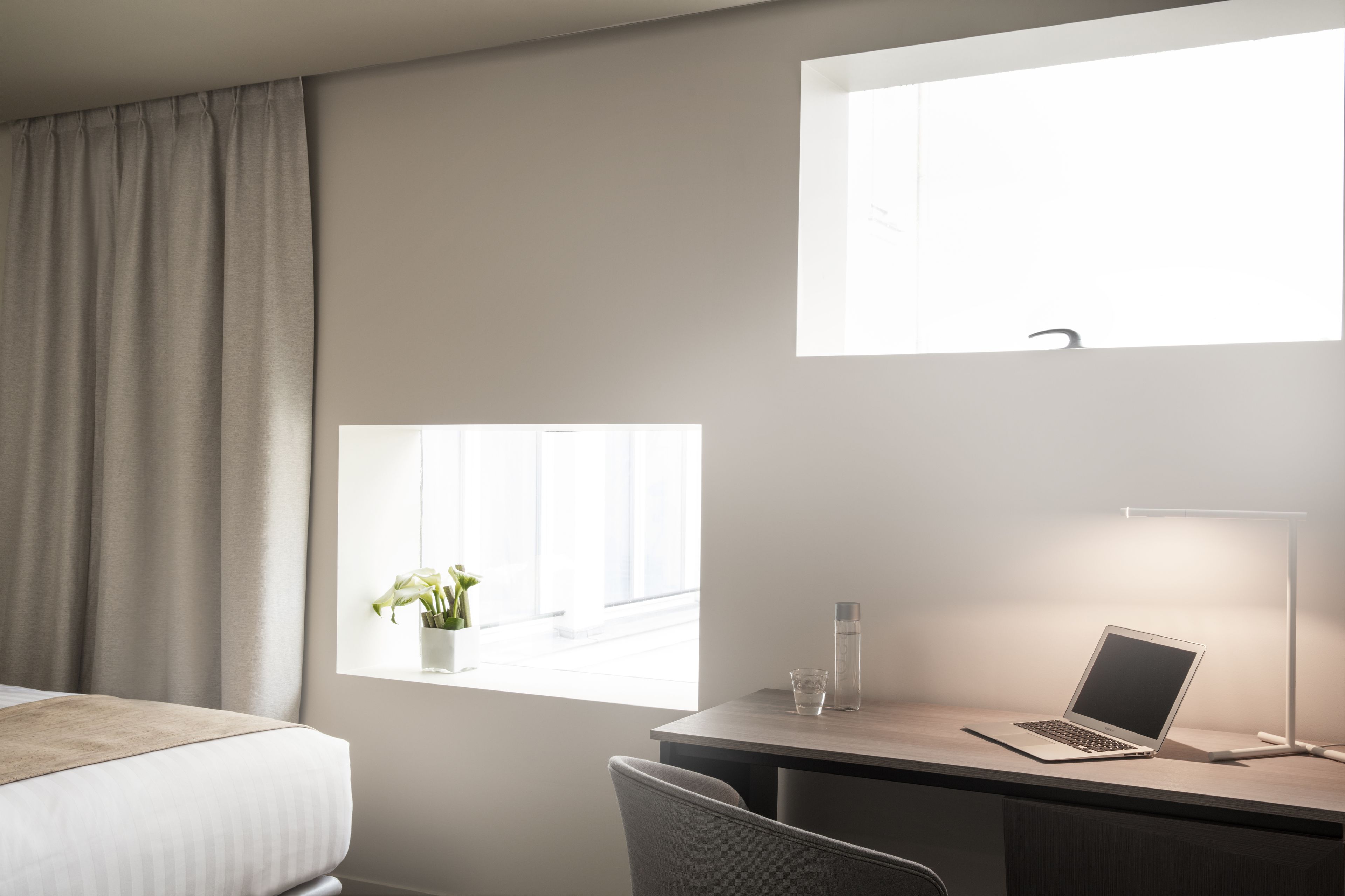 A hotel room with a bed, a desk with a laptop, a desk lamp, a window with a view, and a potted plant.