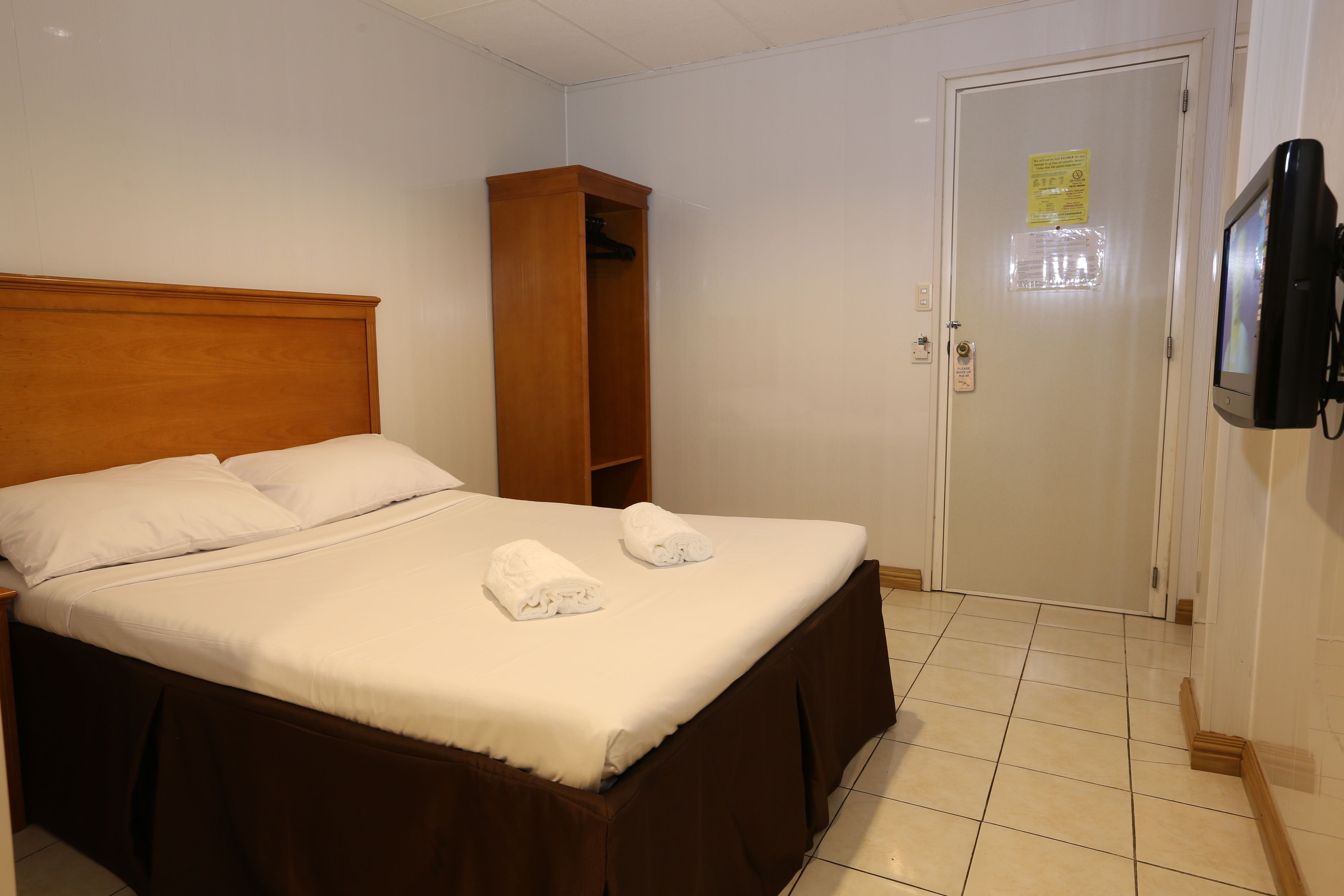 Una cama bien hecha con sábanas blancas y dos toallas, un cabecero de madera, un armario pequeño de madera y una puerta con un cartel.