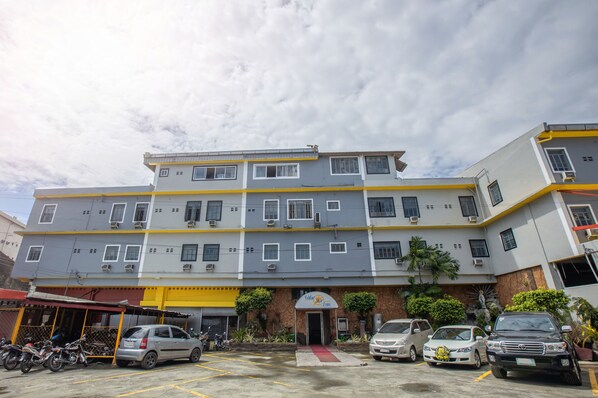 A multi-story building with a yellow and grey facade, multiple windows, and a parking area with several cars and motorcycles.