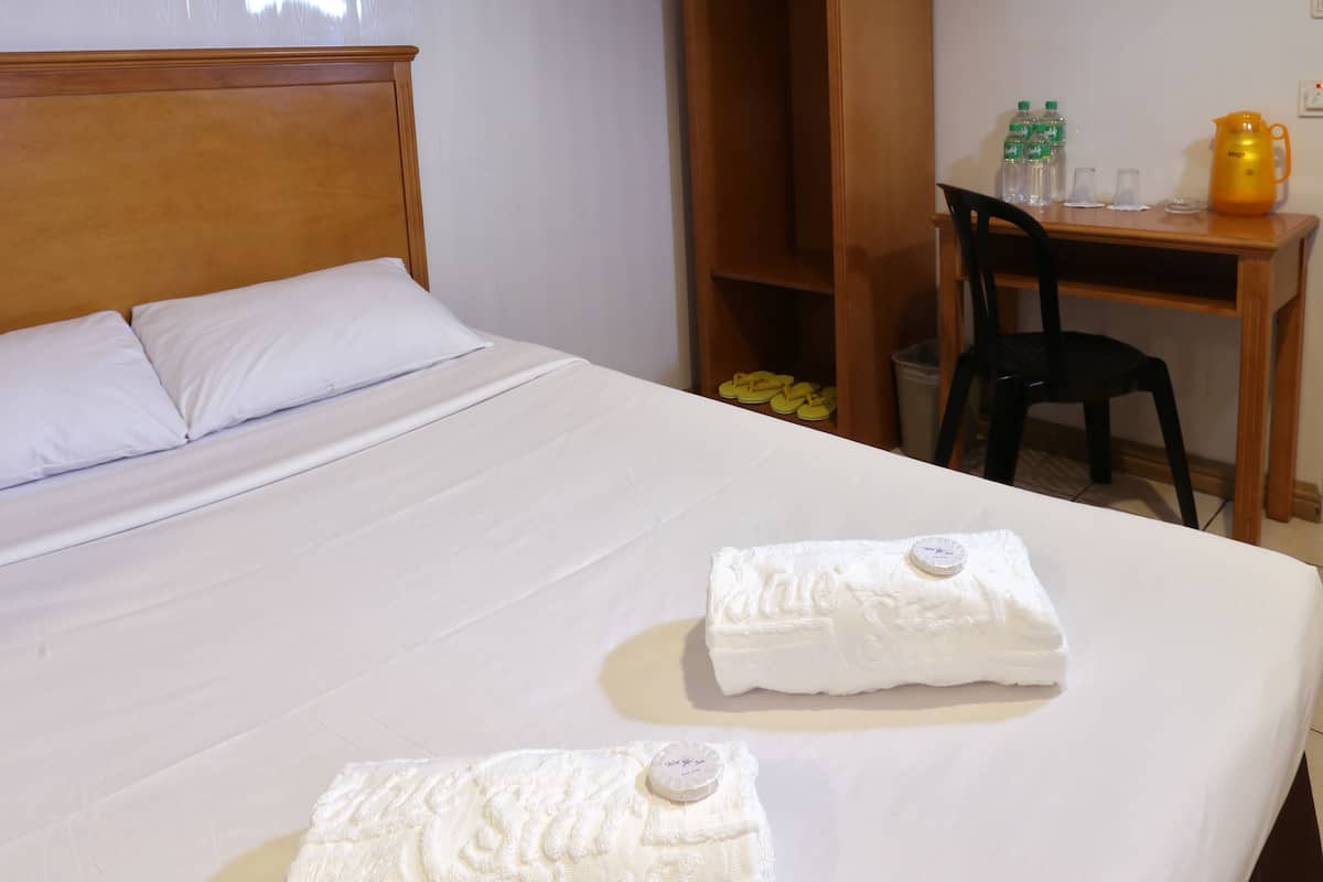 Habitación de hotel con una cama grande, dos toallas, un armario de madera, un escritorio con una silla y una mesita con una tetera y botellas.