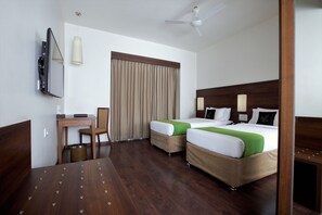 A hotel room with two beds, a desk, a chair, a ceiling fan, and a window with curtains. - Temple Tree Hotel (Bengaluru)