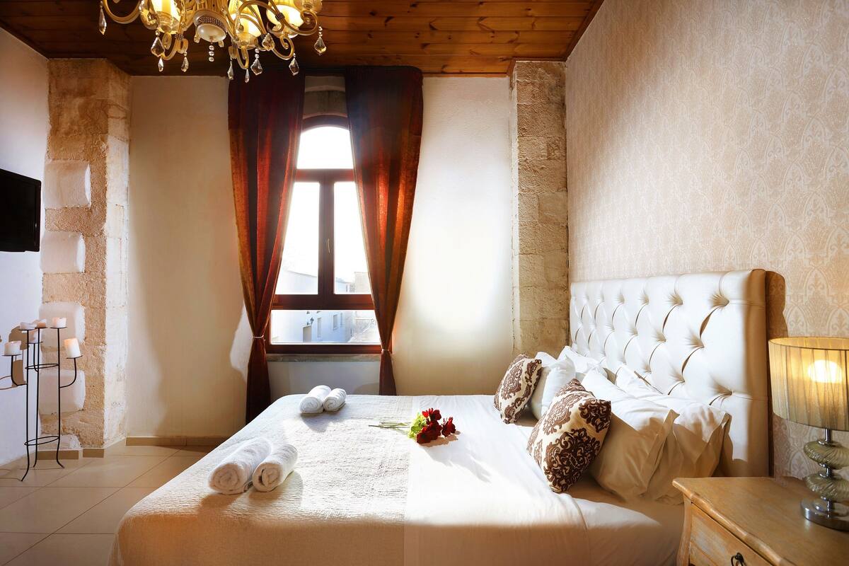 A hotel room with a large bed, a chandelier, a window with curtains, and a wall-mounted TV.