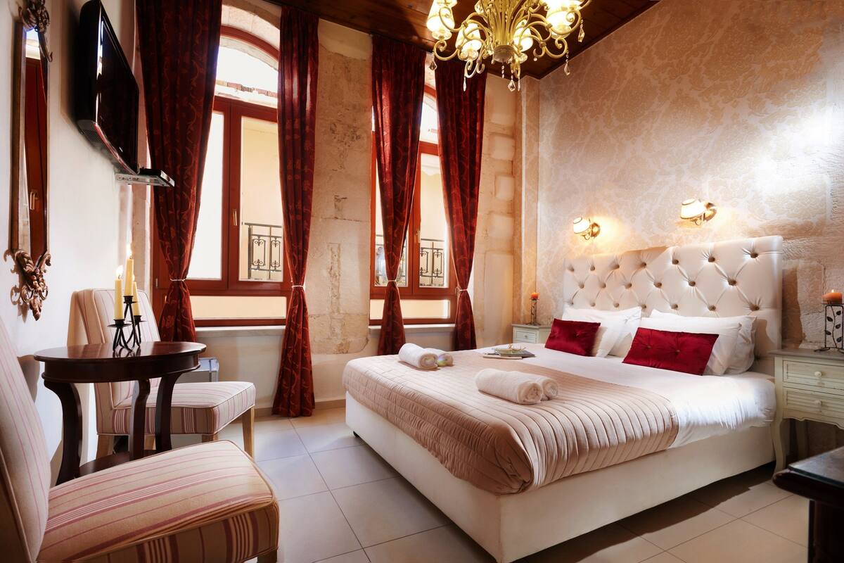 A hotel room with a large bed, a bedside table, a chair, a TV, and a chandelier.