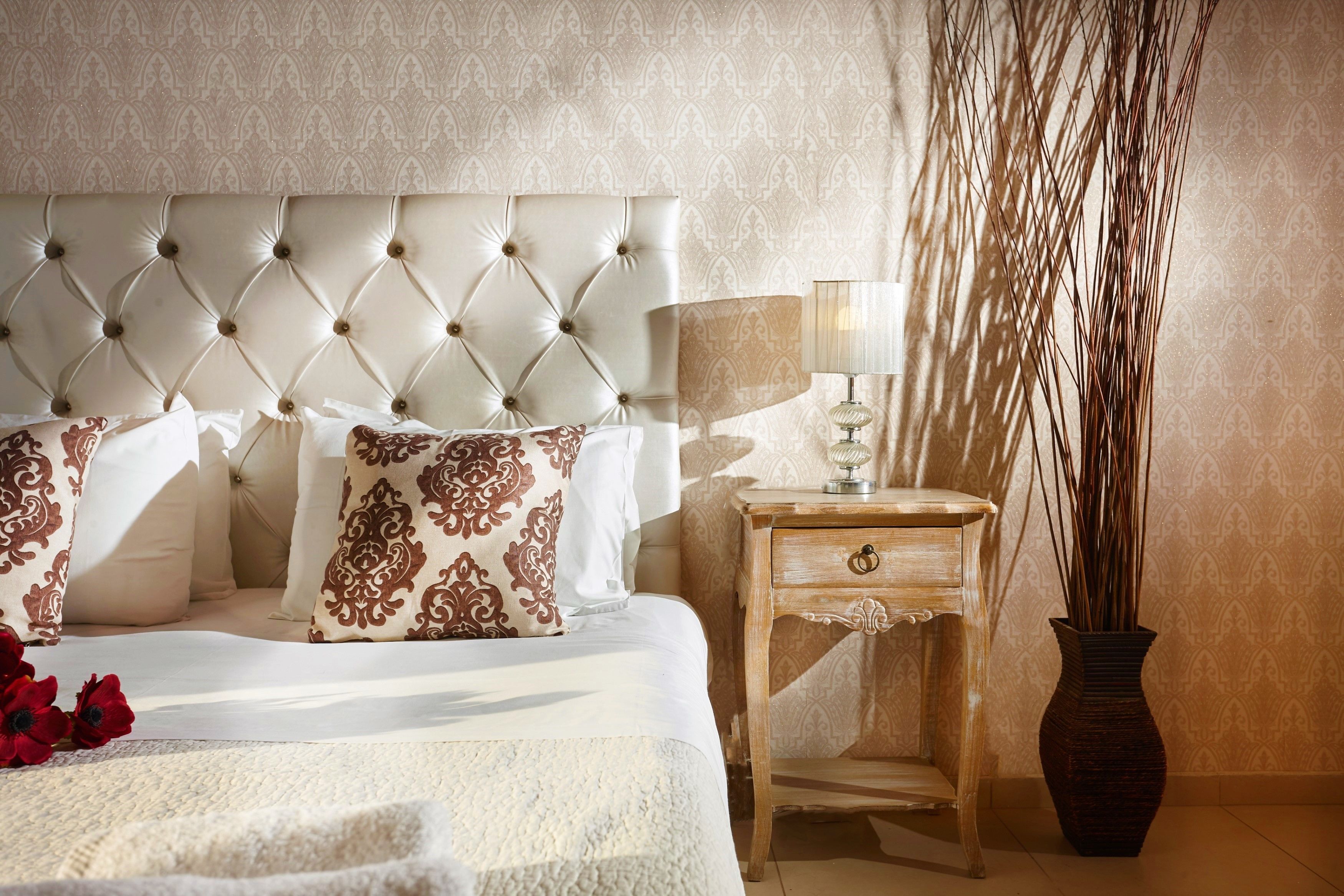 A bedroom with a tufted headboard, a bedside table with a lamp, and a vase with tall plants.