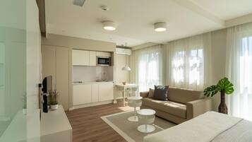 A modern hotel room with a kitchenette, a sofa, a dining table, and a large window with curtains.