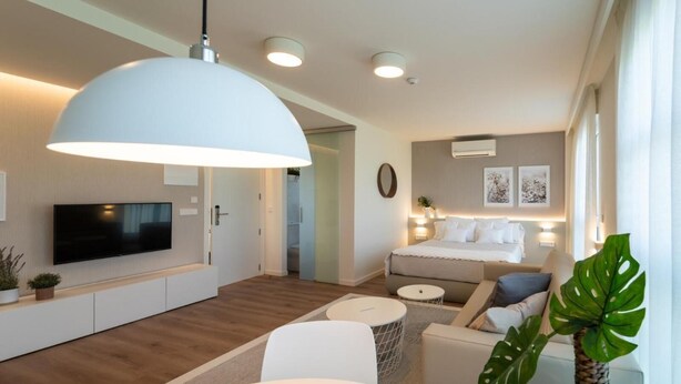 En moderne stue med et fladskærms-tv, en stor hvid pendel, et spiseområde med et rundt bord, og et hyggeligt soveværelse med en seng og et natbord.