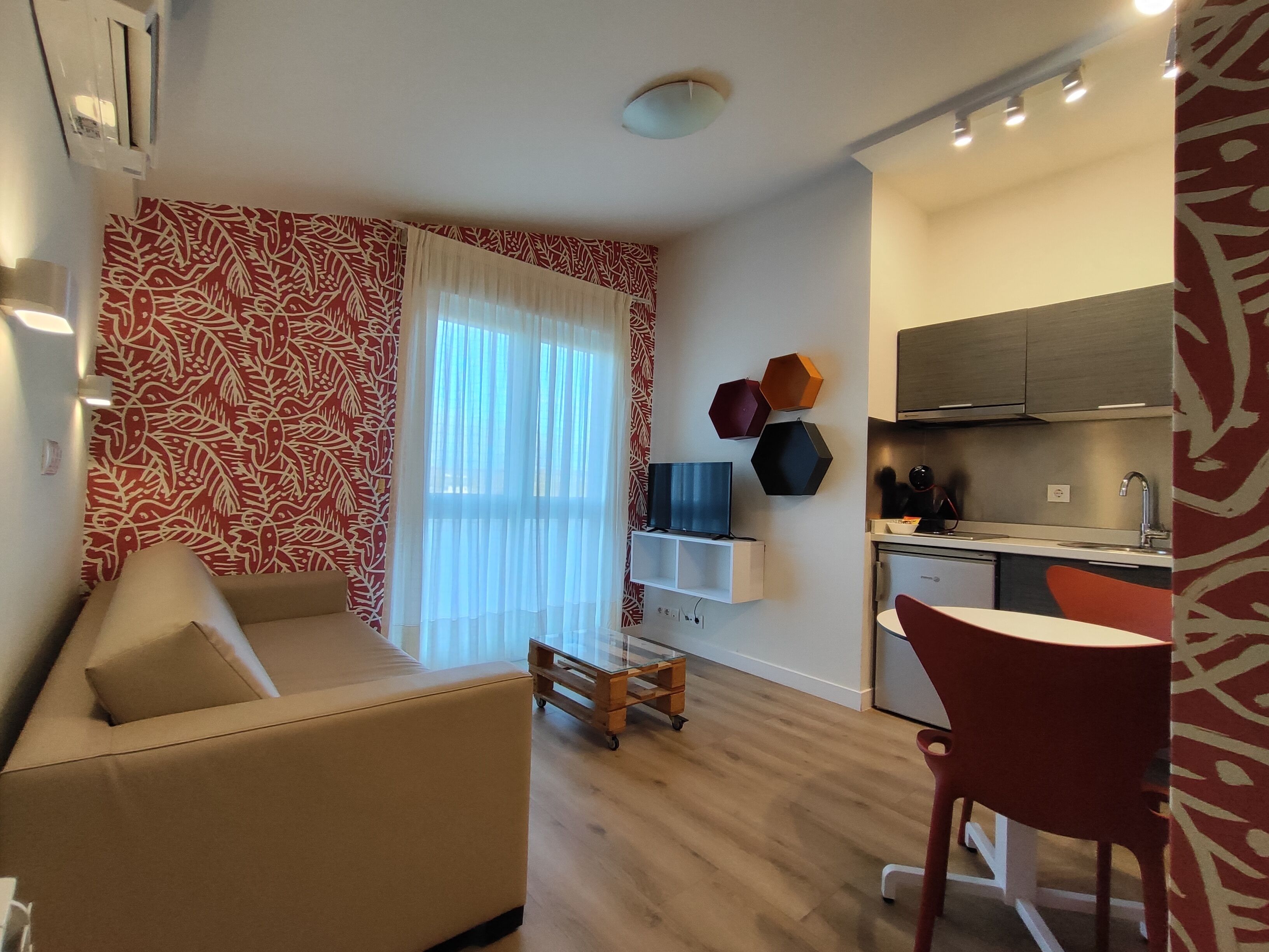 A compact living space with a sofa, a small table, a kitchen area with cabinets and a sink, and a window with sheer curtains.