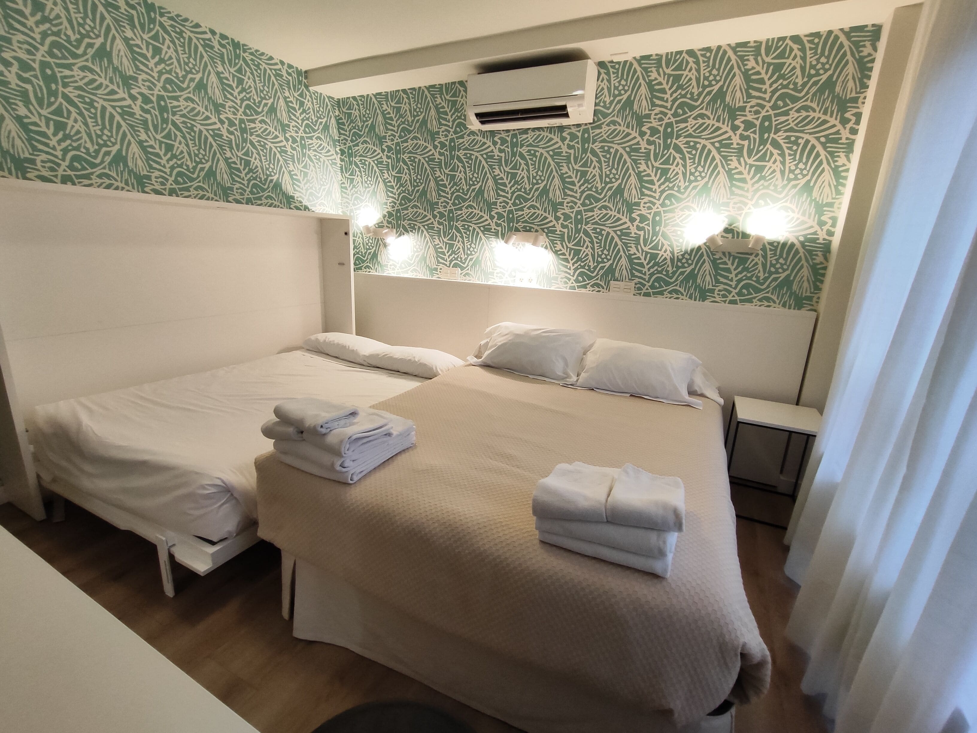 Een hotelkamer met twee bedden, een hoofdbord met ingebouwde verlichting, een split-airco en behang met een patroon.