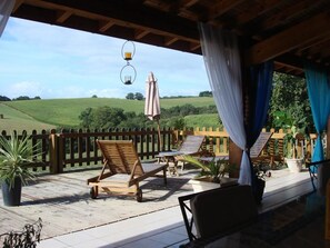 A covered outdoor deck with wooden furniture, a wooden fence, and a view of a green field.