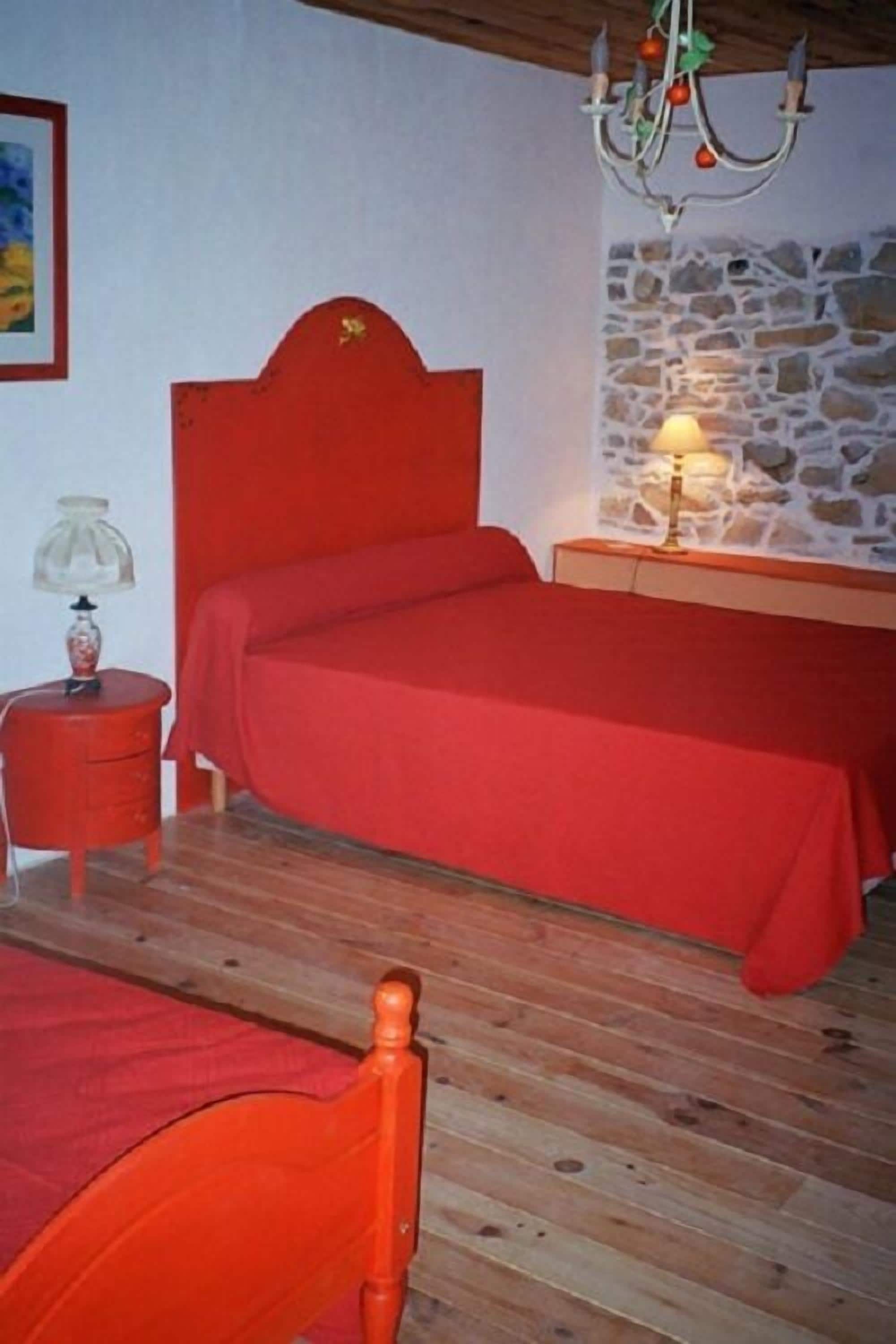 A bedroom with a wooden floor, a red bed, a red chair, a nightstand with a lamp, and a stone wall.