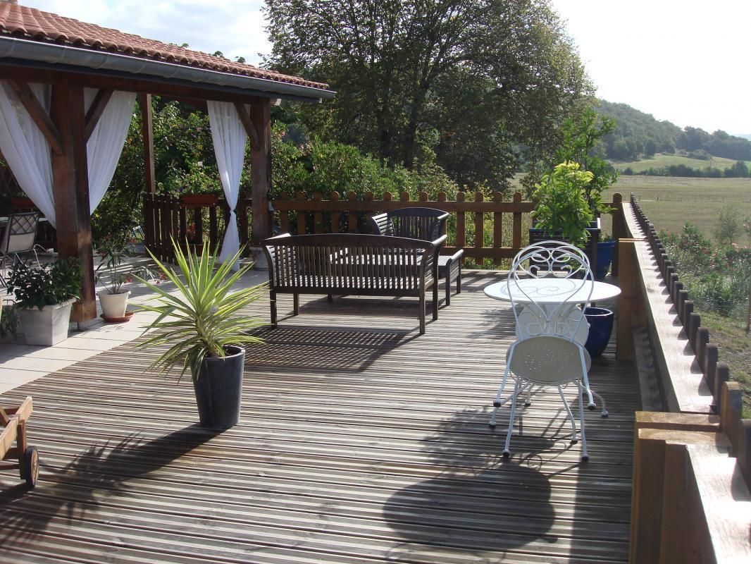 Une terrasse en bois avec un banc, une table et des chaises, entourée de plantes en pot et d’une clôture en bois, d’où l’on peut admirer un paysage verdoyant.