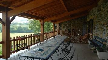 A covered outdoor dining area with a long table and benches, stone walls, and a view of the countryside.