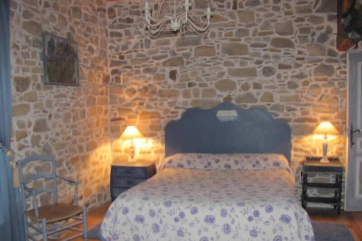 A bedroom with a stone wall, a bed with a floral bedspread, two bedside lamps, a chair, and a wooden floor.
