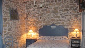 Un dormitorio con pared de piedra, una cama con colcha estampada, dos lámparas de noche, una silla y suelo de madera.