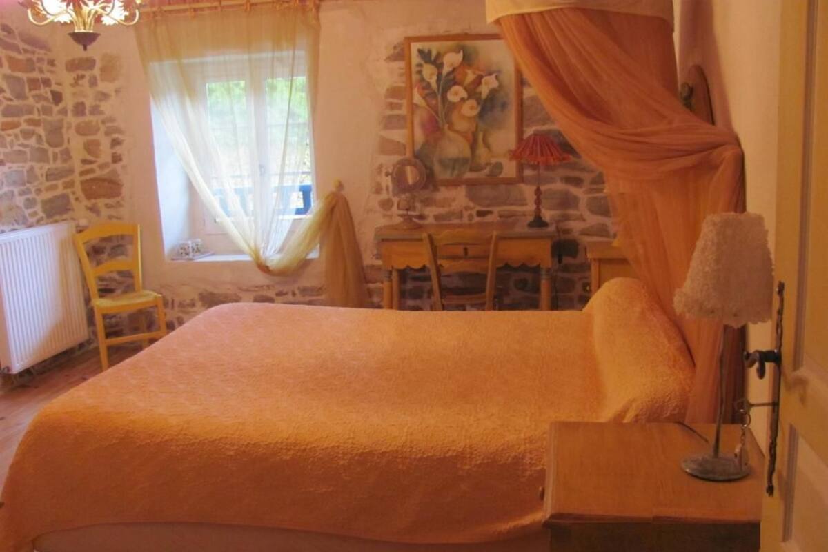 A cozy bedroom with a large bed, a wooden nightstand, a chair, a desk, and a window with curtains.