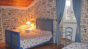 Un dormitorio con pared de piedra, una cama con colcha estampada, una mesita de noche con lámpara, una silla y una ventana con cortinas.