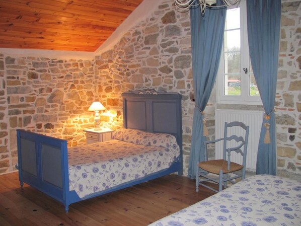 A bedroom with a stone wall, a bed with a floral bedspread, a nightstand with a lamp, a chair, and a window with curtains.