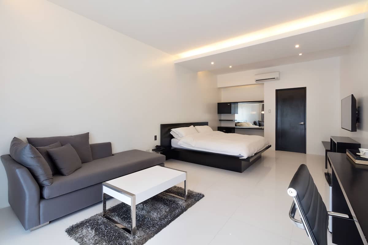 Une chambre d’hôtel moderne avec un canapé gris, un lit blanc, un bureau et un téléviseur à écran plat.
