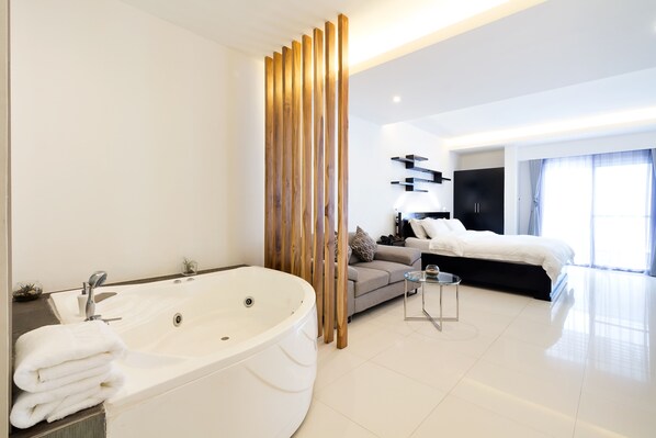 Une chambre d’hôtel moderne avec une grande baignoire blanche, un canapé, une petite table et un lit doté d’une tête de lit noire.