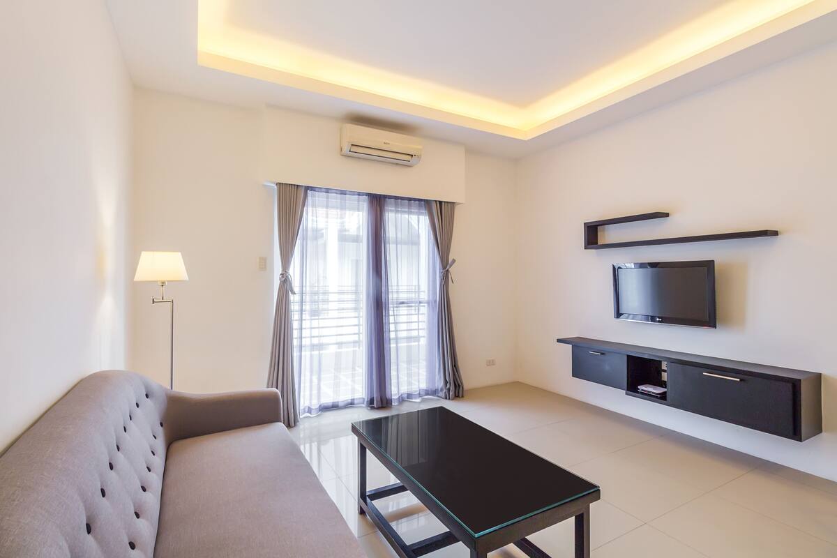 Un salon moderne avec un canapé, une table basse en verre, un téléviseur à écran plat fixé au mur et un lampadaire.
