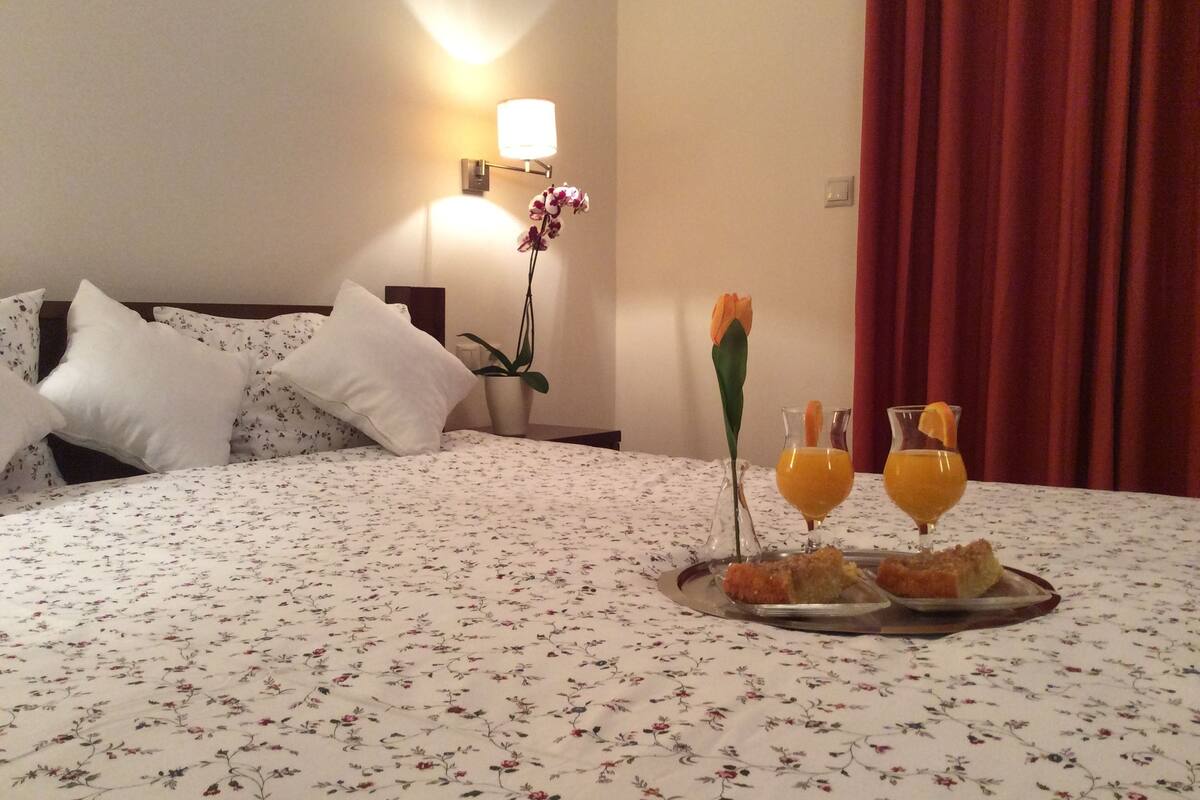 A hotel room with a floral bedspread, a bedside table with a vase of flowers, a tray with orange juice and a rose, and a red curtain.