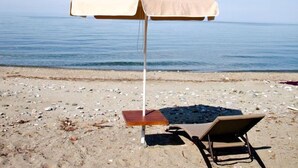 A beach with a single beige umbrella, a wooden table, and a brown lounge chair. - Hotel Morfeas (Dio-Olympos)