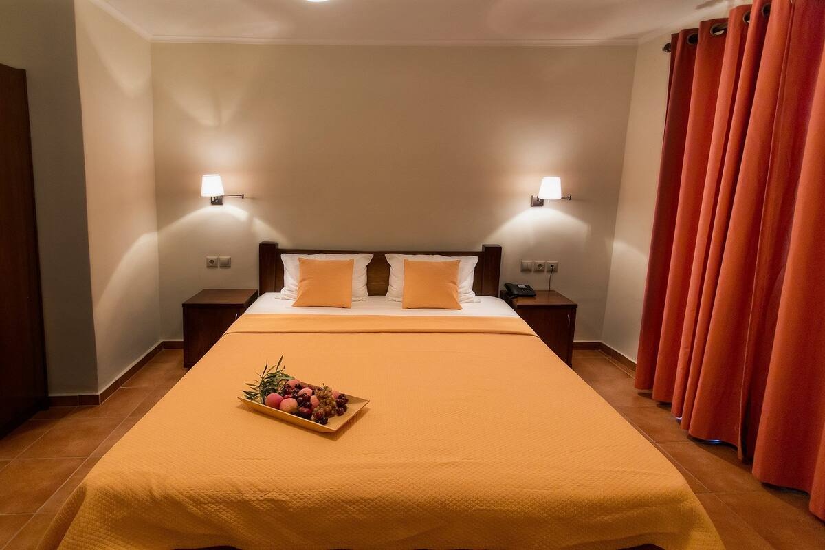 A hotel room with a large bed, two bedside tables, a fruit basket on the bed, and a red curtain.