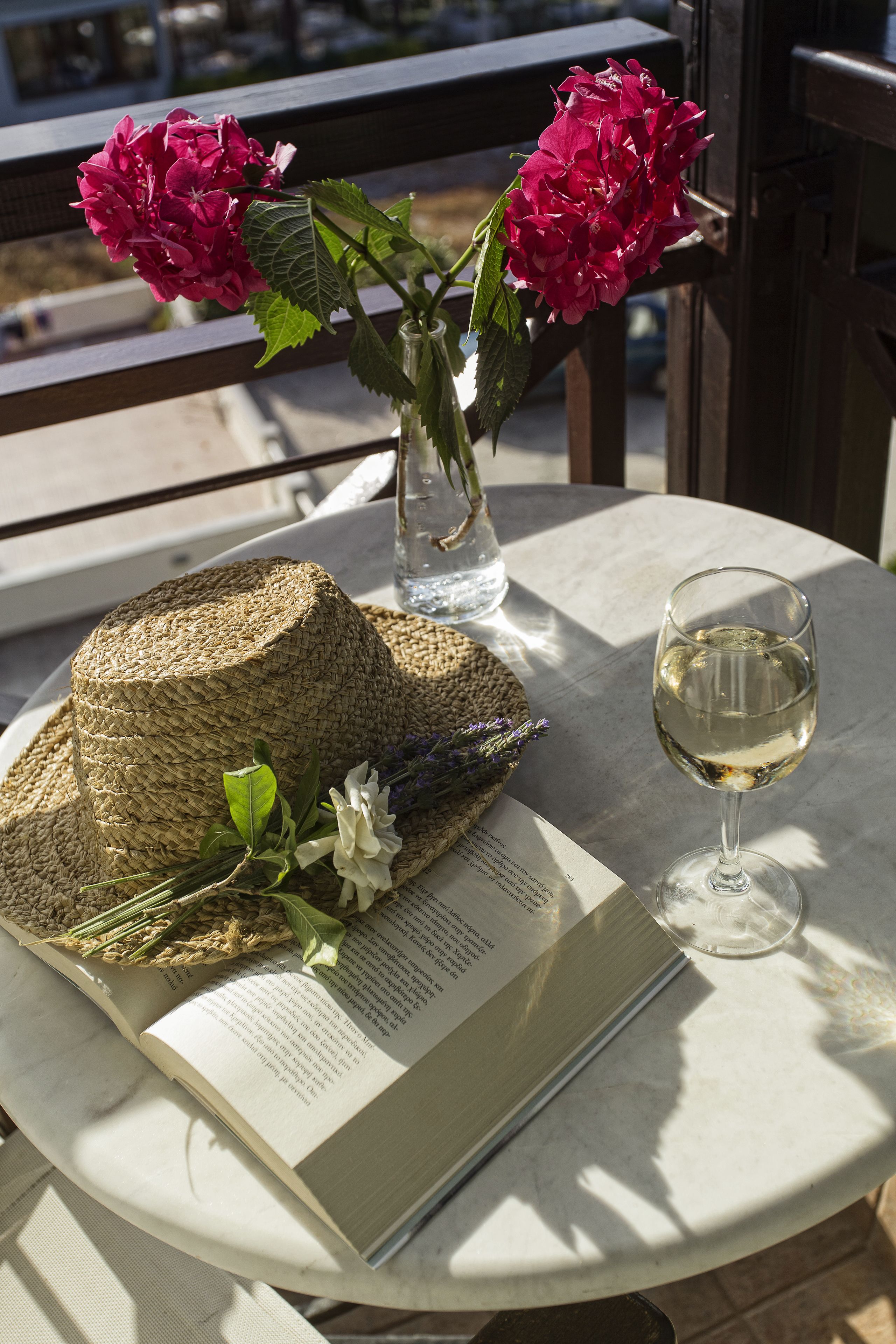 A table with a book, a glass of white wine, a straw hat, and a vase with flowers.