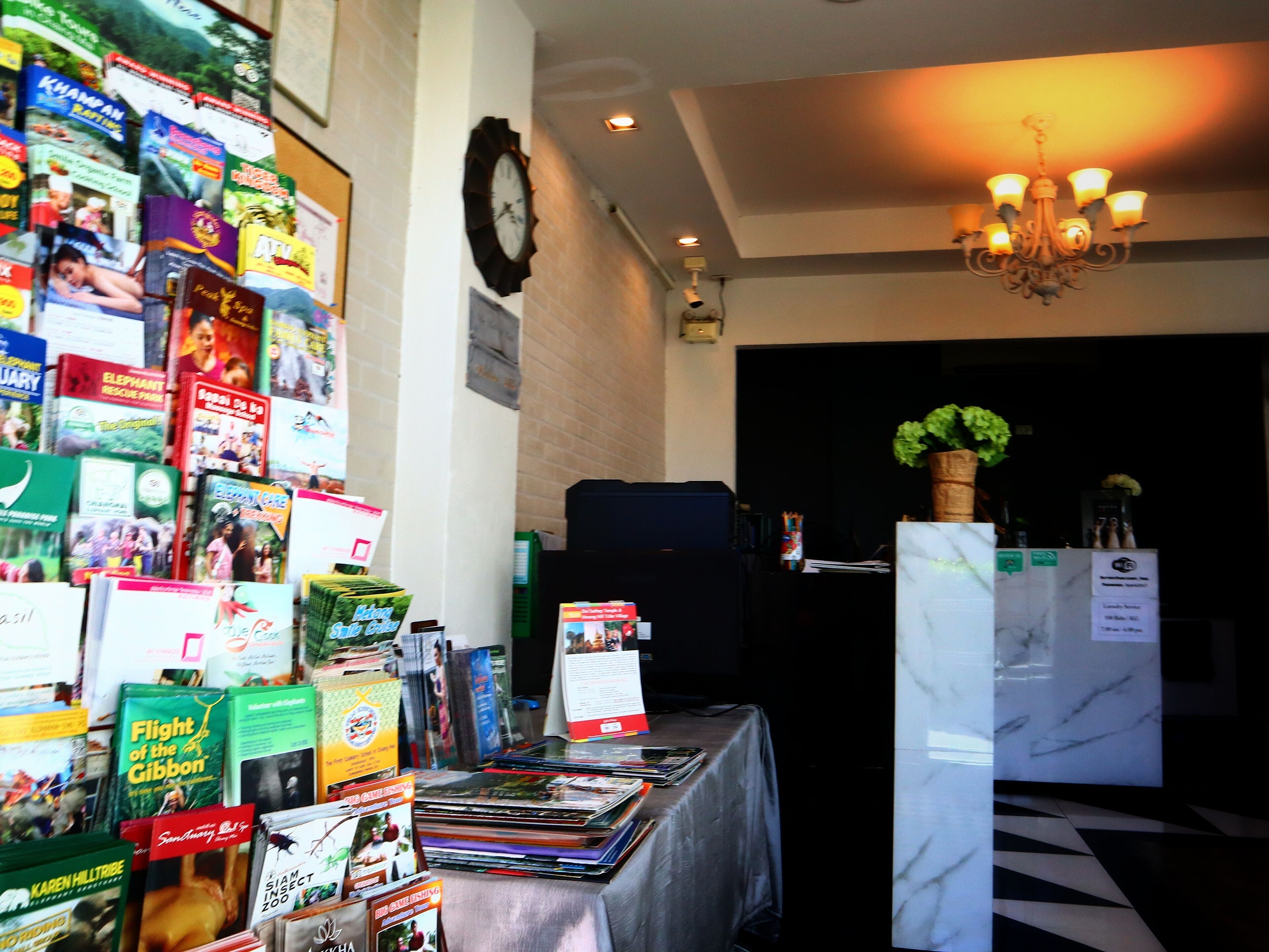 A room with a table displaying various books and magazines, a clock on the wall, and a chandelier.