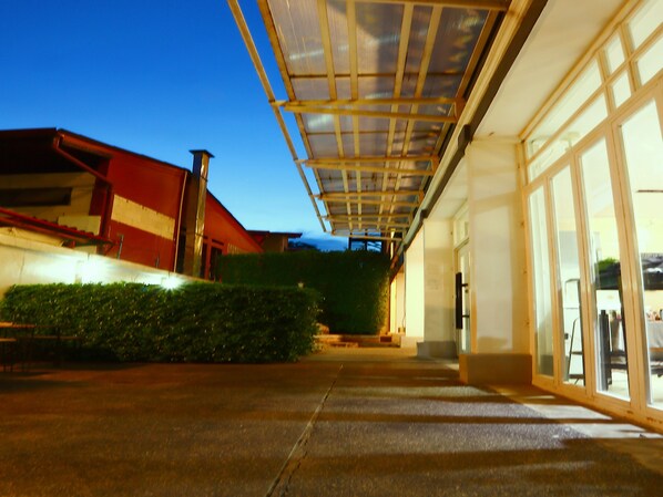 Un espacio al aire libre cubierto con una puerta de vidrio que da a un edificio, un seto y una estructura de techo rojo al fondo.