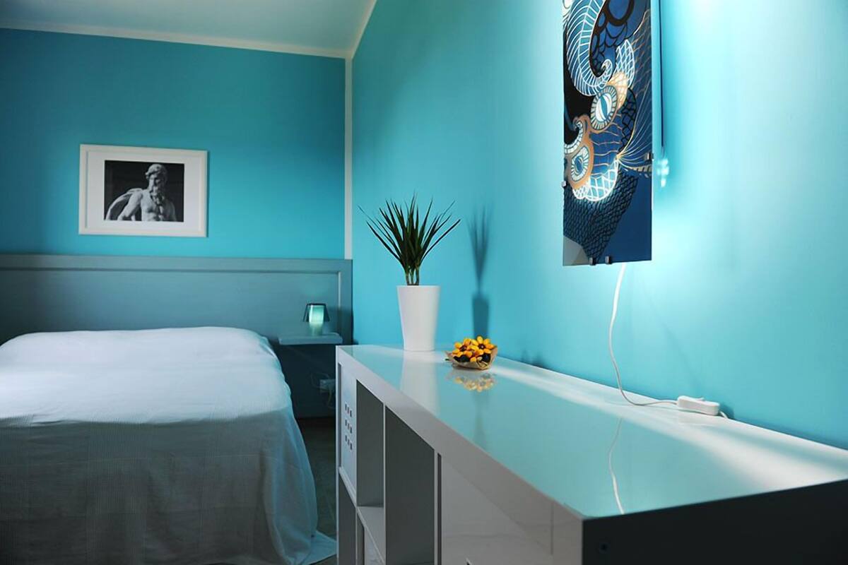 A bedroom with a bed, a nightstand, a white vase with a plant, and a wall-mounted artwork.