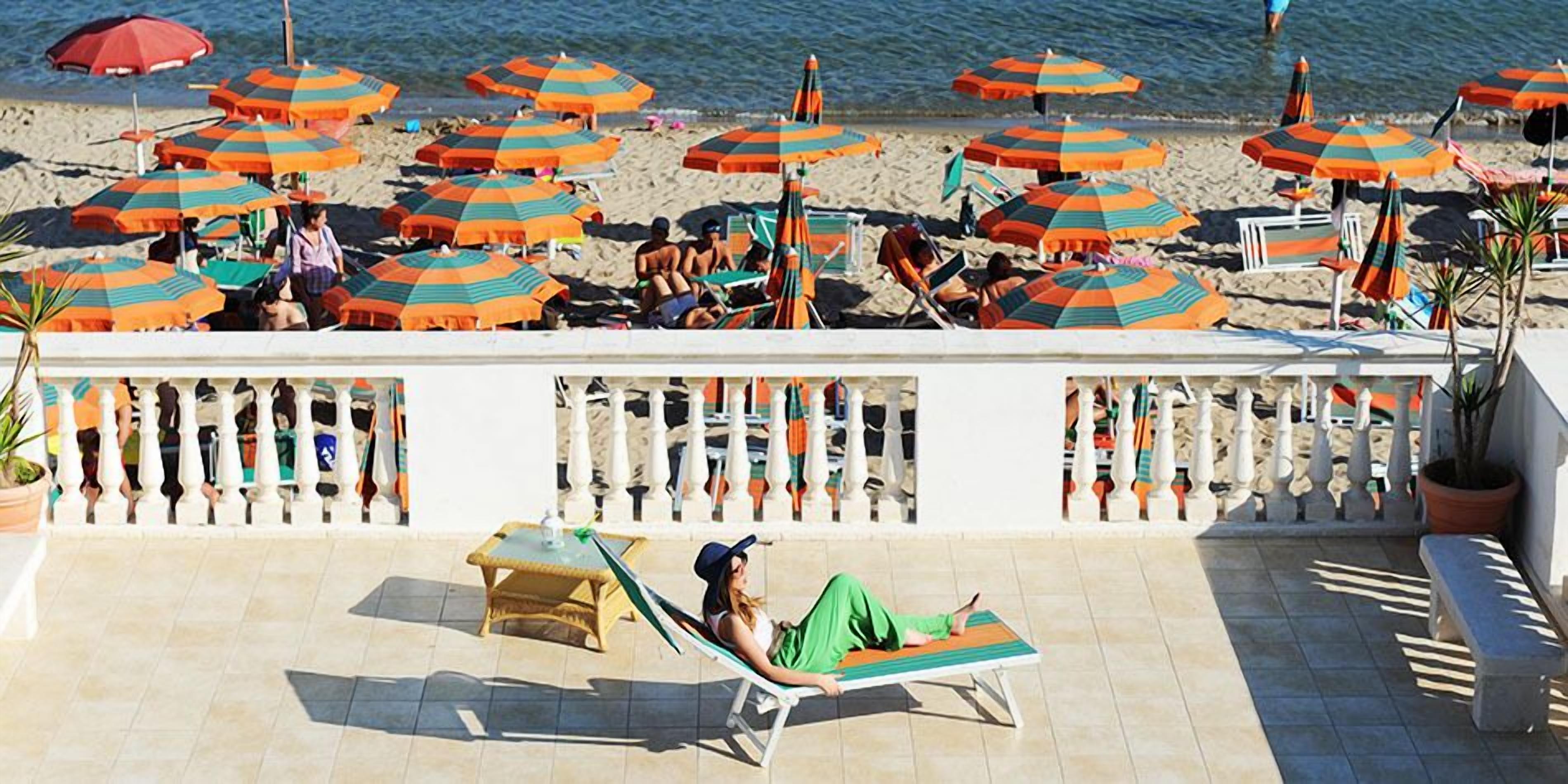 Un área de descanso junto a la playa con varios sillones de sol y sombrillas, una barandilla y plantas en macetas.