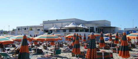 A beach with numerous striped umbrellas and lounge chairs, a building in the background, and clear skies.