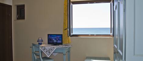 A room with a bed, a desk with a television, a window with a view of the sea, and a door.