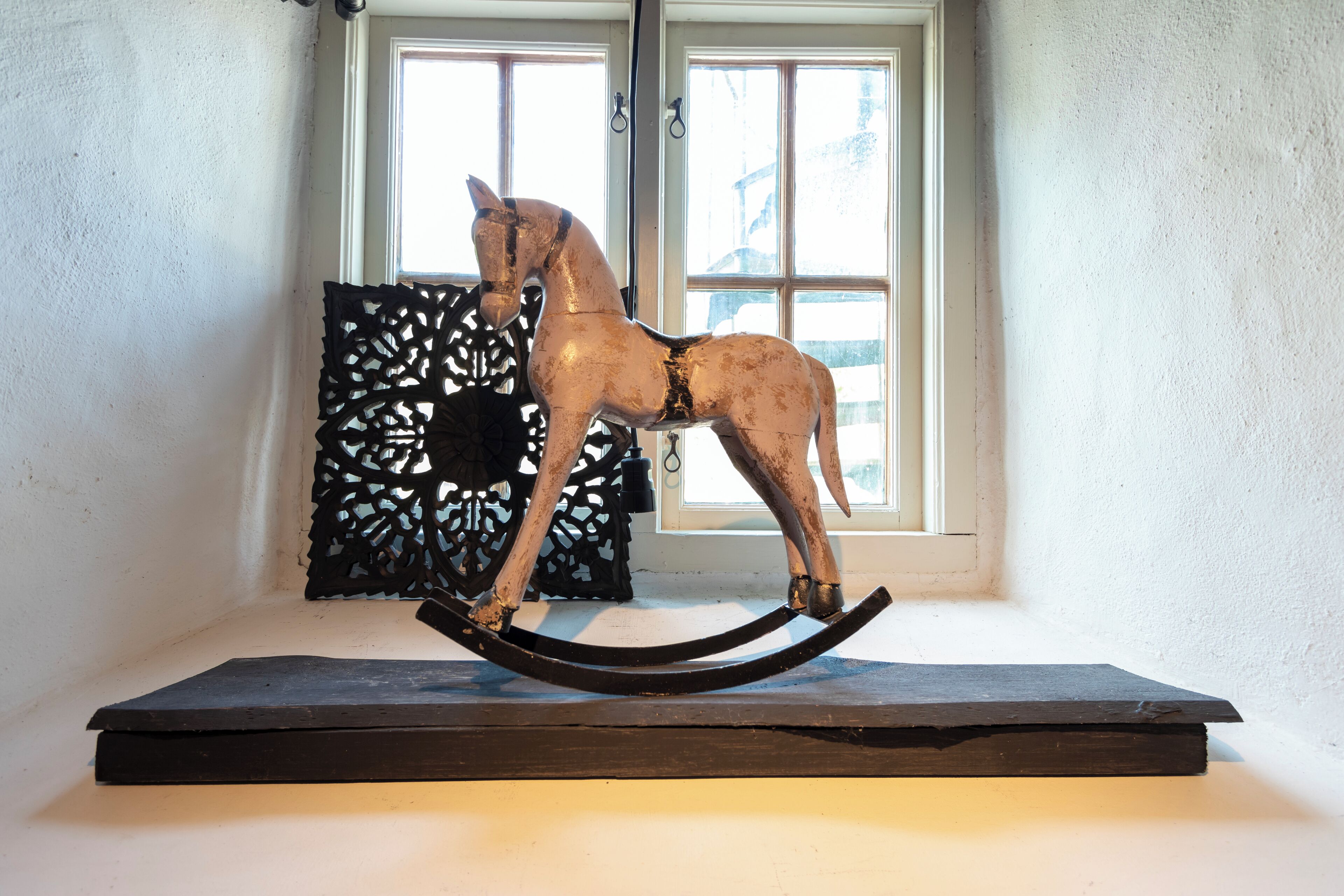 Una escultura de caballo balancín sobre una plataforma de madera oscura en una habitación con paredes blancas y un ventanal.
